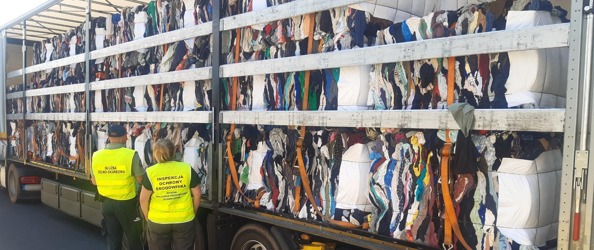 Dwie osoby w kamizelkach odblaskowych stoją przy otwartej naczepie samochodu ciężarowego wypełnionej sprasowanymi belami odpadów tekstylnych; ładunek jest zabezpieczony pasami transportowymi i ułożony na całej długości pojazdu.