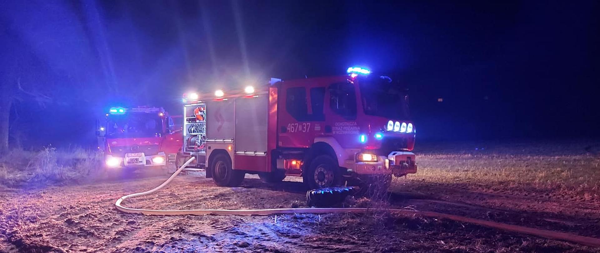 Zdjęcie przedstawia dwa pojazdy ciężarowe straży pożarnej na polu w porze nocnej.do jednego podłączony jest wąż strażacki.Oba wozy maja włączone sygnały.