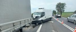 Widać uszkodzony samochód dostawczy, po lewej naczepa samochodu ciężarowego, po prawej przejeżdżające pojazdy 