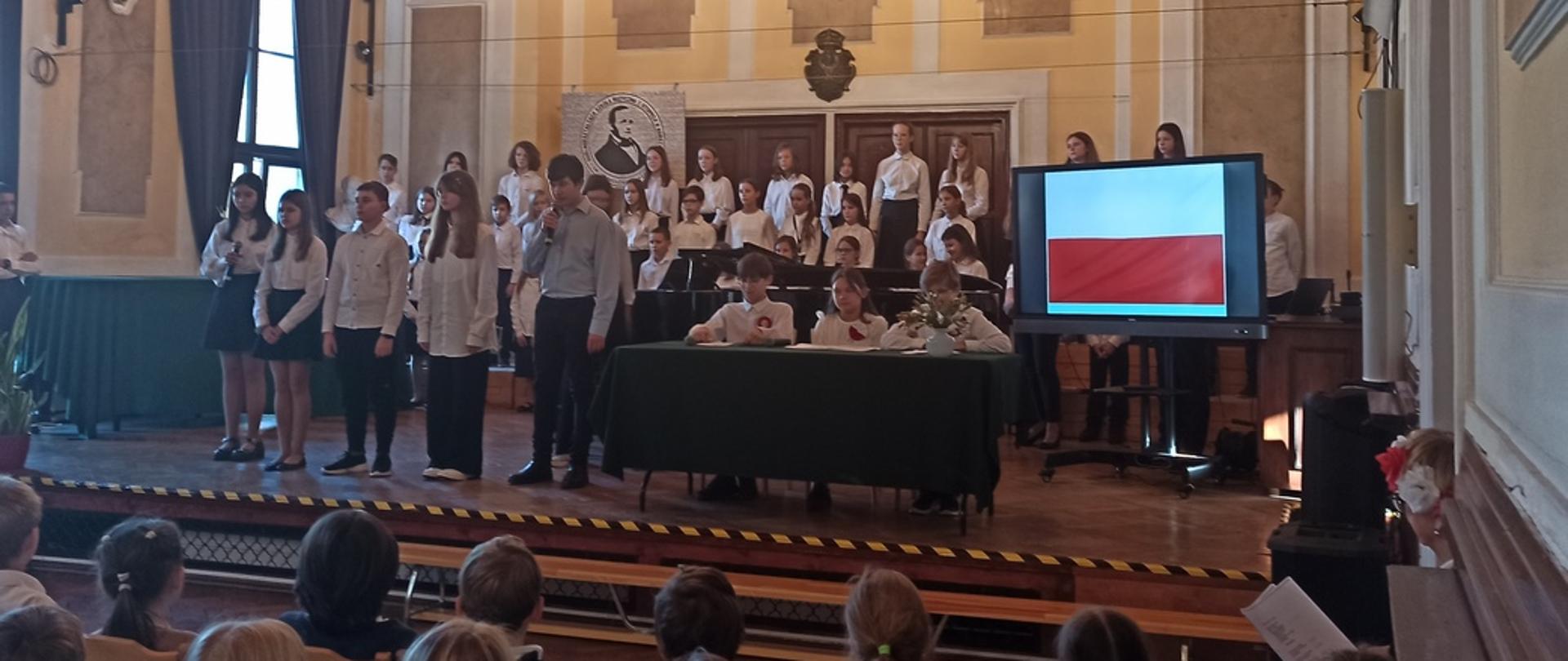 Na scenie chór i uczniowie klasy 7, bliżej widać dzieci oglądające akademię