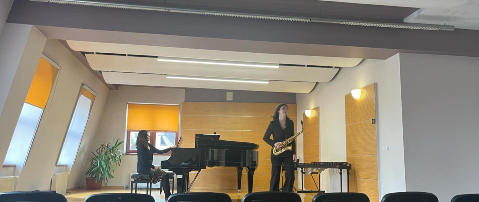 Zdjęcie przedstawia scenę w dużej sali koncertowej, prawdopodobnie w konserwatorium lub akademii muzycznej, takiej jak Akademia Muzyczna im. Fryderyka Chopina w Warszawie. Na scenie znajdują się dwie osoby i instrumenty muzyczne. Po lewej stronie, z profilu, siedzi osoba grająca na czarnym fortepianie. Po prawej stronie, stojąc, znajduje się osoba grająca na saksofonie. Obie osoby wydają się być elegancko ubrane, prawdopodobnie w ciemne stroje. Za pianistką widać zieloną roślinę doniczkową. Na tle sceny, za muzykami, znajduje się ściana w jasnym, drewnianym kolorze, z kilkoma panelami dźwiękochłonnymi. Po obu stronach sceny widać wysokie okna, przez które wpada naturalne światło. Okna mają skośne górne krawędzie, dopasowane do kształtu sufitu. Przed sceną, w dolnej części zdjęcia, widać rzędy pustych, czarnych krzeseł, ustawionych dla publiczności. Oświetlenie w sali jest mieszane, naturalne światło z okien uzupełniają lampy sufitowe i kinkiety ścienne, które tworzą przyjemną atmosferę. 
