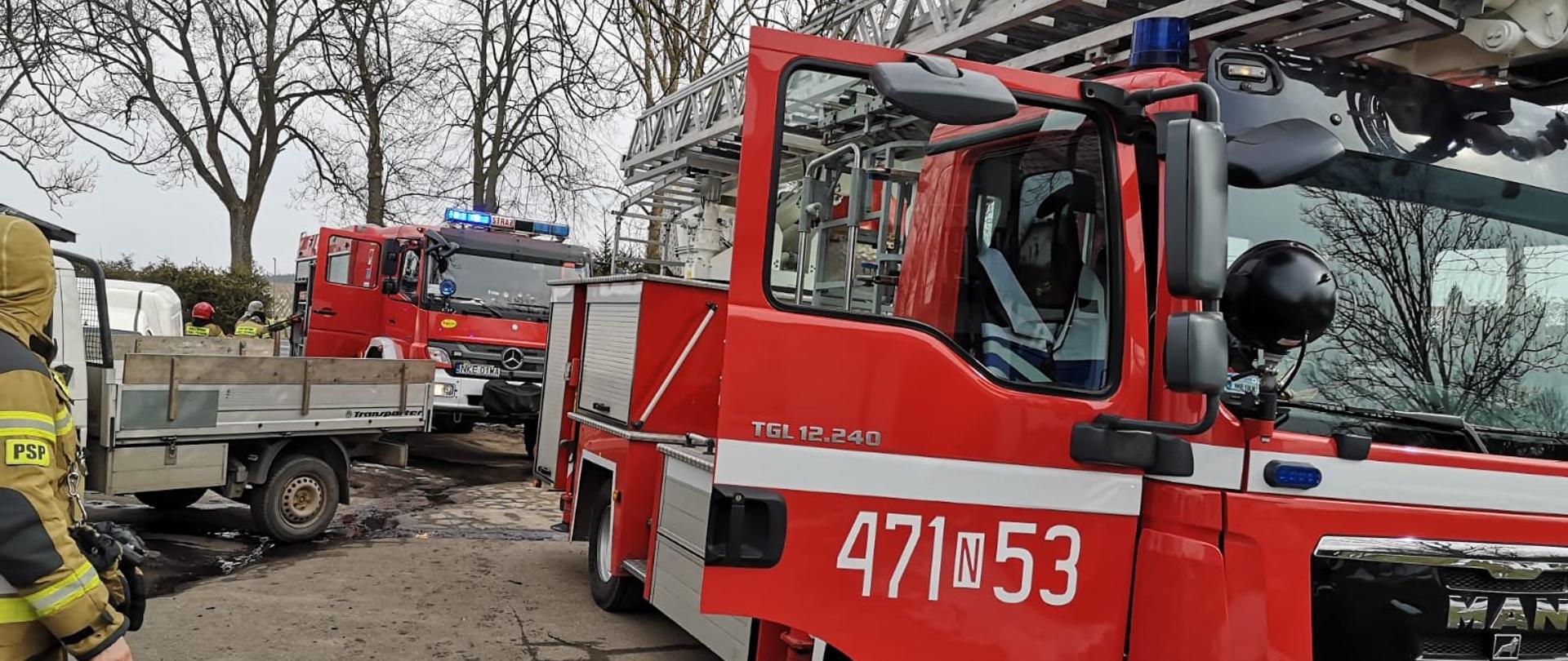 Zdjęcie przedstawia dwa czerwone wozy strażackie ustawione na drodze przed budynkiem w którym doszło do pożaru oraz biały samochód dostawczy zakładu stolarskiego. Strażacy w piaskowych ubraniach bojowych wyciągają sprzęt z samochodu.