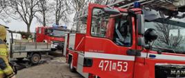 Zdjęcie przedstawia dwa czerwone wozy strażackie ustawione na drodze przed budynkiem w którym doszło do pożaru oraz biały samochód dostawczy zakładu stolarskiego. Strażacy w piaskowych ubraniach bojowych wyciągają sprzęt z samochodu.