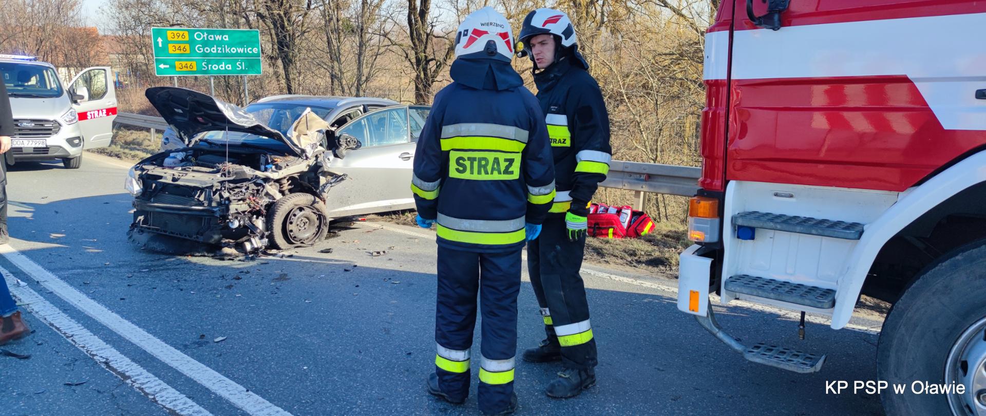 na zdjęciu widać rozbity samochód osobowy uczestniczący w wypadku. Po prawej dwaj strażacy rozmawiają ze sobą.