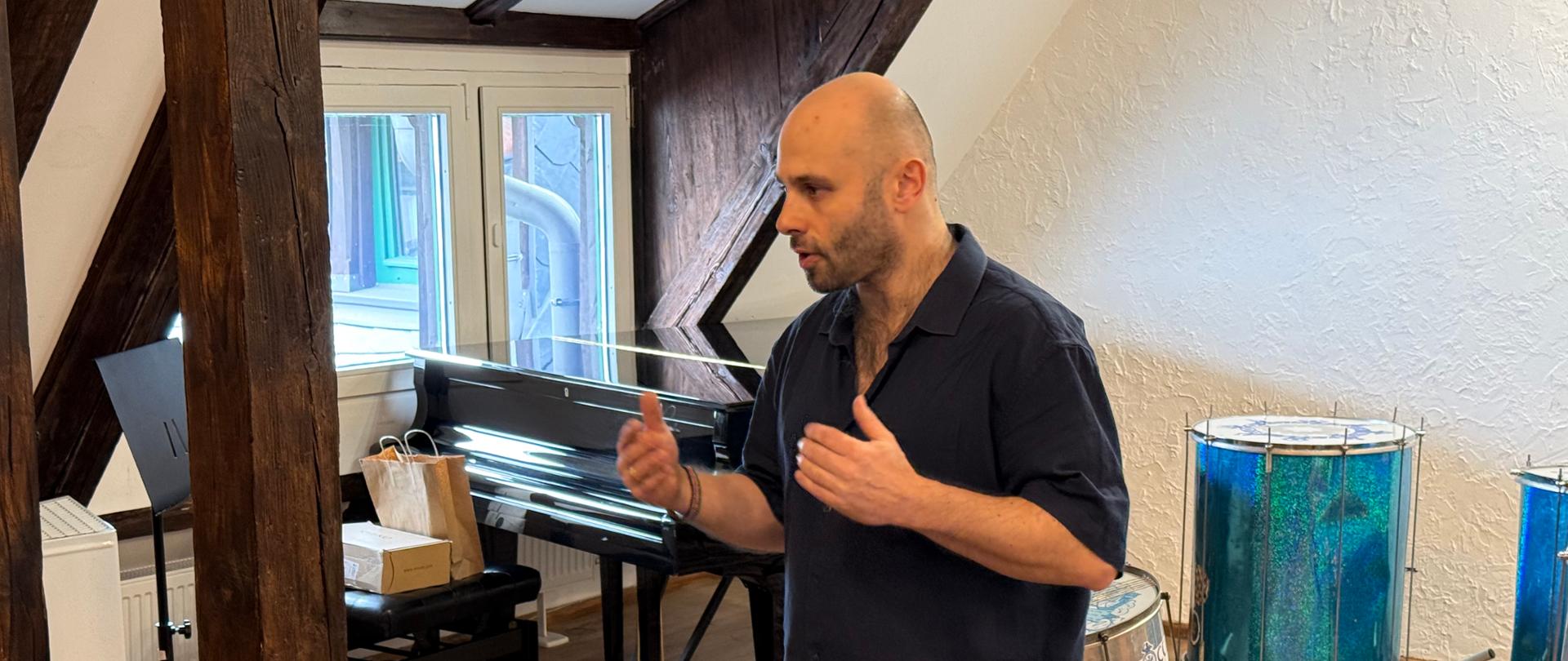 Mężczyzna stojący w jasnym pomieszczeniu z drewnianymi belkami, gestykulujący podczas mówienia. W tle widoczny fortepian oraz zestaw instrumentów perkusyjnych, w tym duże bębny ustawione na stojakach i otwarta walizka z pałkami