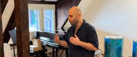 Mężczyzna stojący w jasnym pomieszczeniu z drewnianymi belkami, gestykulujący podczas mówienia. W tle widoczny fortepian oraz zestaw instrumentów perkusyjnych, w tym duże bębny.
