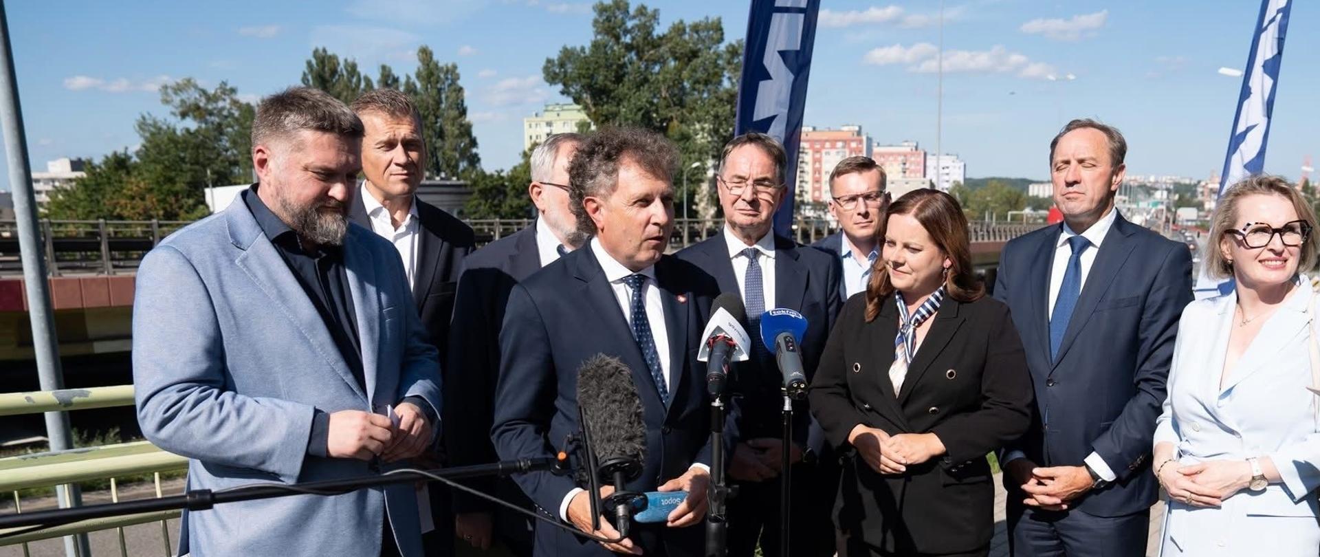 Dziewięć osób stoi obok siebie. Wśród nich mówiący do mikrofonów wiceminister Jacek Karnowski.