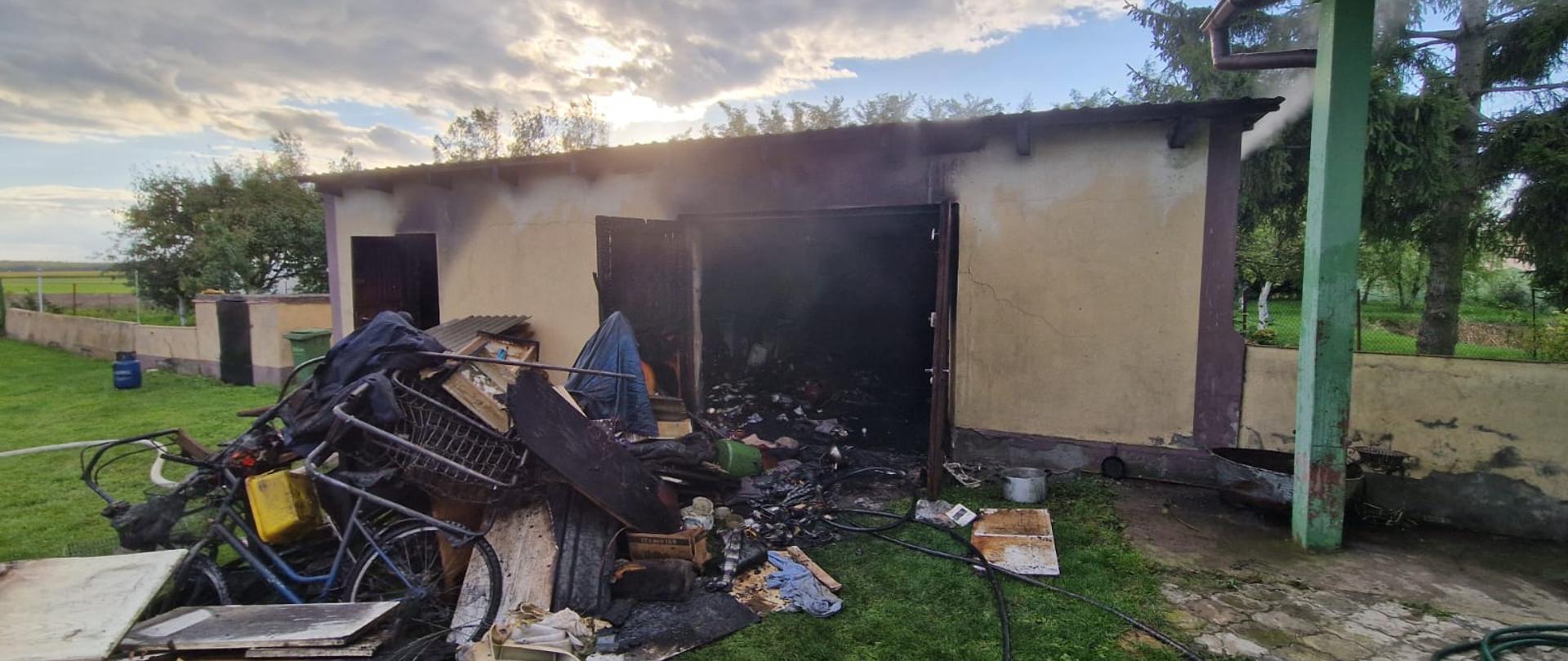 Na zdjęciu widoczny jest budynek gospodarczy z częściowo okopconą elewacją i otwartymi, nadpalonymi drzwiami. Z wnętrza wydobywa się ciemny dym. Przed budynkiem leży sterta nadpalonych i wyniesionych z środka przedmiotów, m.in. elementy drewniane, metalowe oraz sprzęty codziennego użytku. Na ziemi rozciągnięte są węże strażackie, a wokół widać trawnik, ogrodzenie i drzewa.