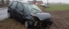 Zdjęcie przedstawia samochód osobowy ze zniszczonym przodem i przednią szybą znajdujący się poza jezdnią. W oddali drugi samochód poza jezdnią ze zniszczonymi lewymi drzwiami. Obok wóz strażacki