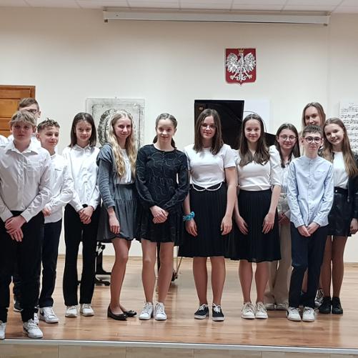 Fotografia przedstawia uczestników audycji fortepianu dodatkowego. Wszyscy stoją na scenie auli PSM I st. w Grajewie. W tle fortepian, grafiki przedstawiające partytury i postać Jana Sebastian Bacha, oraz fragment drzwi w kolorze brązowym. 