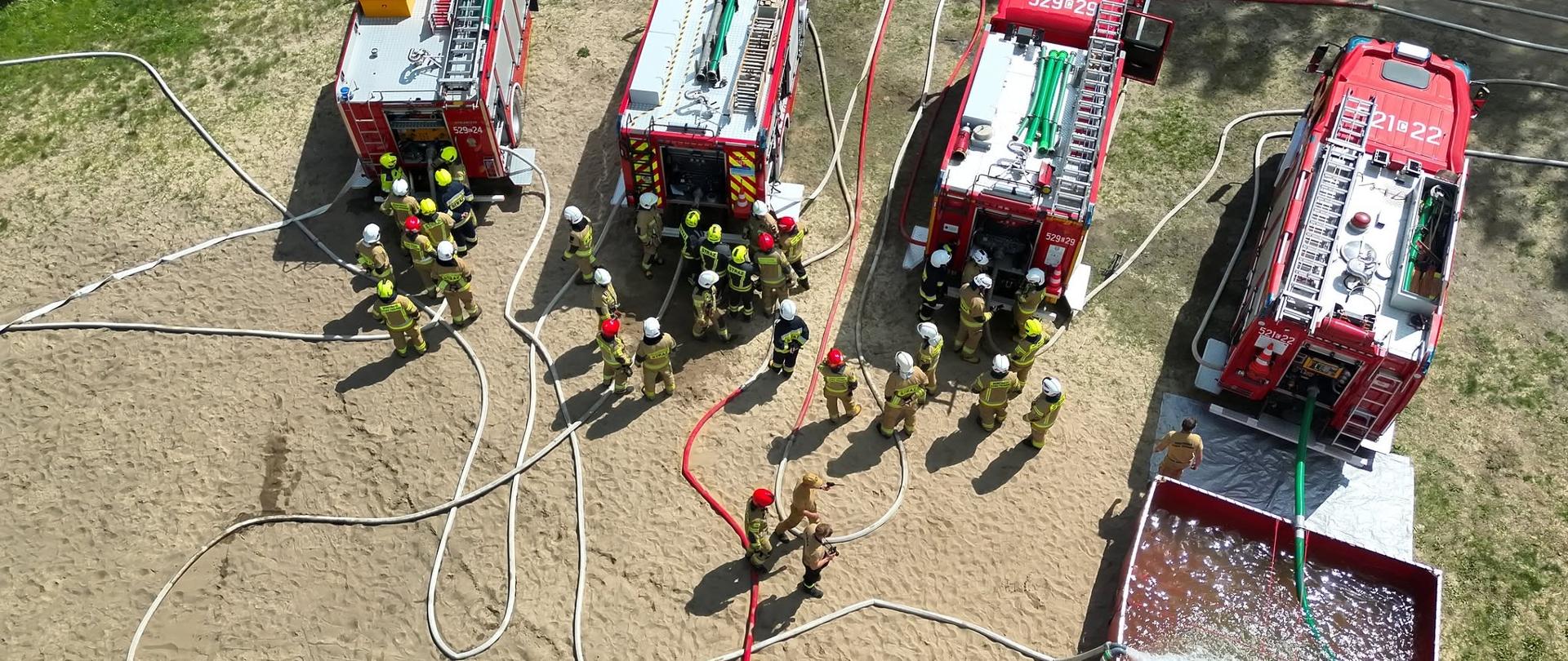 Zdjęcie przedstawia widok z góry na ćwiczenia strażackie zorganizowane na piaszczystym terenie. W rzędzie stoi pięć czerwonych wozów bojowych, z których cztery są ustawione tyłem, a od ich autopomp rozchodzą się liczne węże pożarnicze. Na dachach pojazdów widoczne są drabiny, węże ssawne oraz numery operacyjne, takie jak 529-24, 529-28 czy 529-29. Wokół samochodów gromadzi się grupa kilkunastu strażaków w charakterystycznych piaskowych ubraniach specjalnych i hełmach. W prawym dolnym rogu zdjęcia umieszczono duży, rozkładany, czerwony zbiornik na wodę, do którego wpada strumień wody pod ciśnieniem, a gruba zielona rura odprowadza ją z powrotem. Całość sceny dopełniają rozciągnięte chaotycznie węże tłoczne, linie zasilające oraz fragmenty zielonej trawy widoczne na obrzeżach piaszczystego placu.