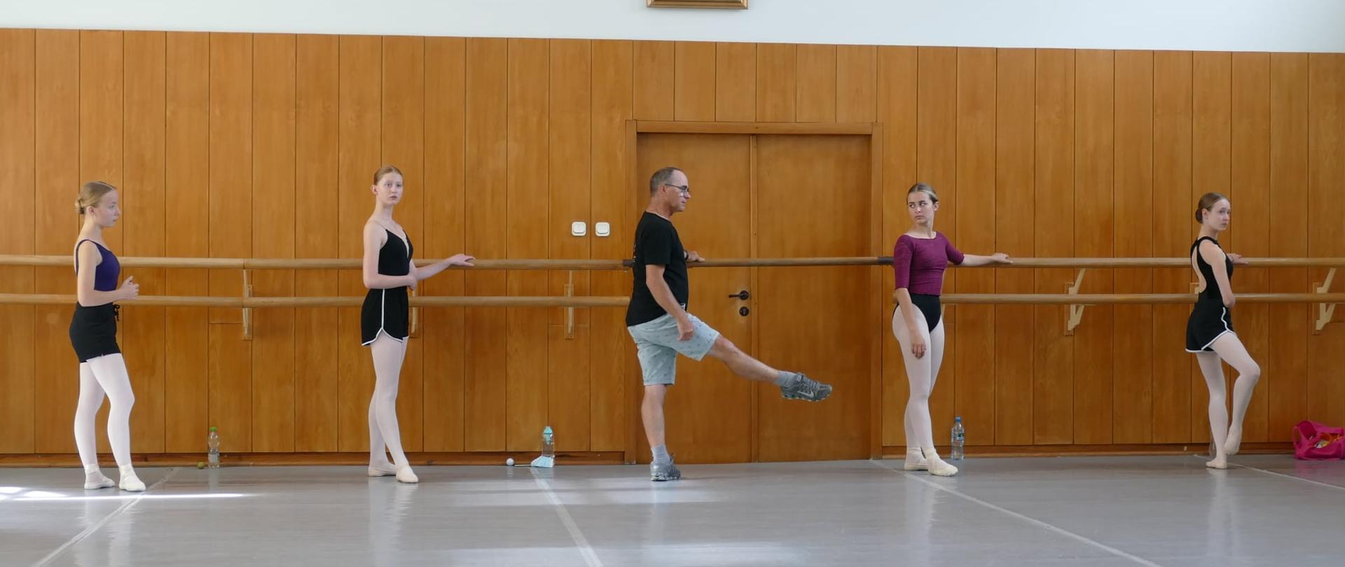 Uczennice stojące przy drążku a po środku nauczyciel znajdują się na sali baletowej 