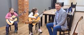 Dwie uczennice siedzą trzymając gitary, obok nauczyciel prowadzący warsztaty.