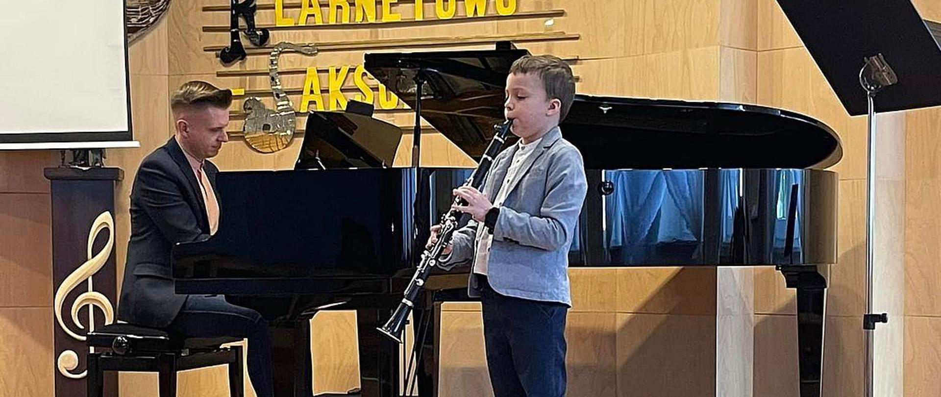Zdjęcie przedstawia ucznia Miłosza Matyszczaka grającego na klarnecie oraz akompaniującego mu na fortepianie Pana Tomasza Ratajczaka podczas występu na scenie w Krośnie Odrzańskim.