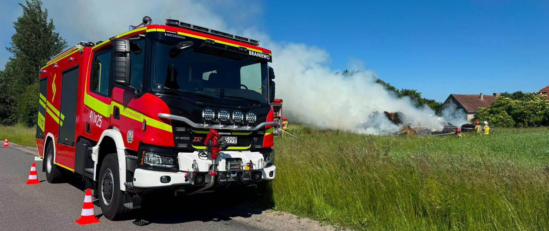 Na zdjęciu duży czerwony samochód strażacki stoi na drodze. Z prawej strony widać pożar, dużo białego dymu. Strażacy gaszą pożar. Błękitne niebo, słonecznie.
