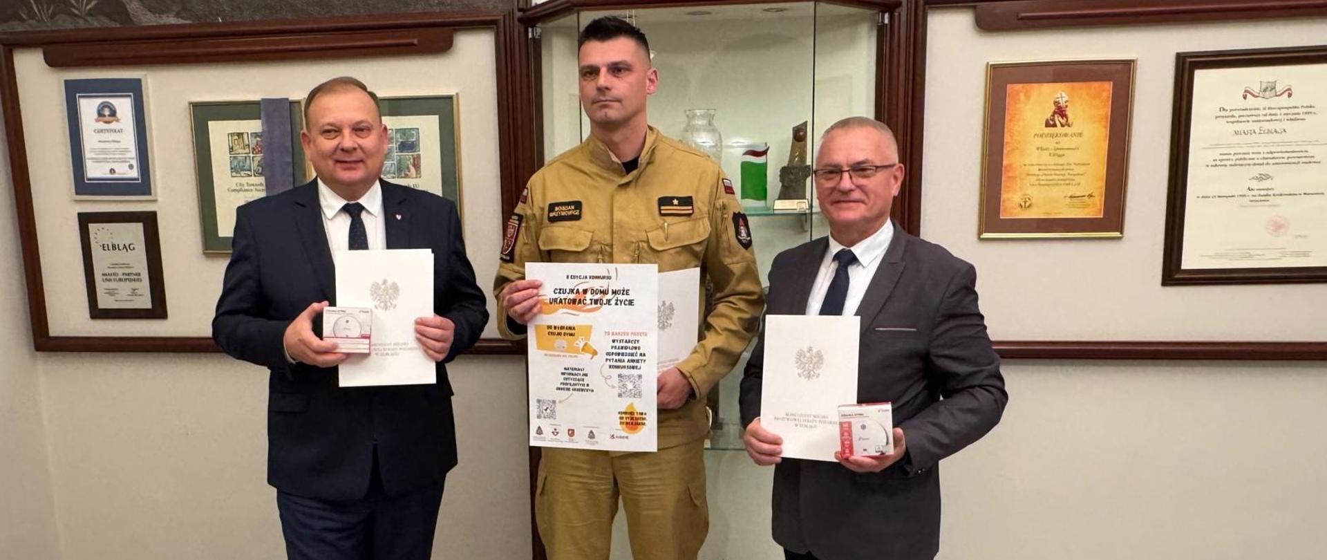 Na zdjęciu znajdują się 3 mężczyźni: Prezydent miasta Elbląga, Wicestarosta elbląski i Komendant Miejski PSP w Elblągu. Mężczyźni stoją w holu Urzędu Miasta w Elblągu, w rękach trzymają podpisany regulamin II edycji konkursu Czujka w domu może uratować Twoje życie oraz plakat promujący to wydarzenie.