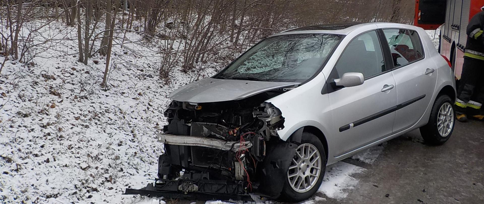 Na zdjęciu widać uszkodzony pojazd Renault Clio znajdujący się na poboczu jezdni. Ma on uszkodzony przód pojazdu i wyrwany przedni zderzak. Przed samochodem leży fragment z uszkodzonego przodu pojazdu. Po prawej stronie zdjęcia widać samochód pożarniczy z włączonymi sygnałami świetlnymi oraz strażaka stojącego przy samochodzie. W tle znajdują się drzewa.