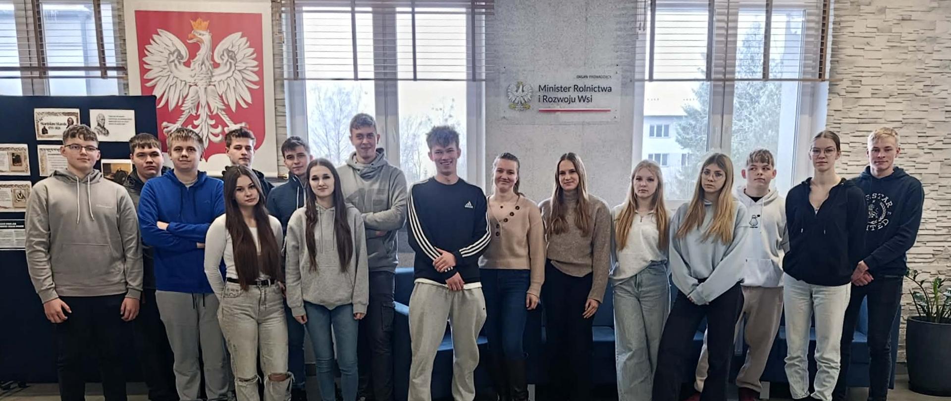Grupa młodzieży stoi w rzędzie, pozując do pamiątkowego zdjęcia w szkolnym holu. W tle widoczne jest godło Polski, tablica informacyjna oraz napis wskazujący na Ministerstwo Rolnictwa i Rozwoju Wsi jako organ prowadzący.