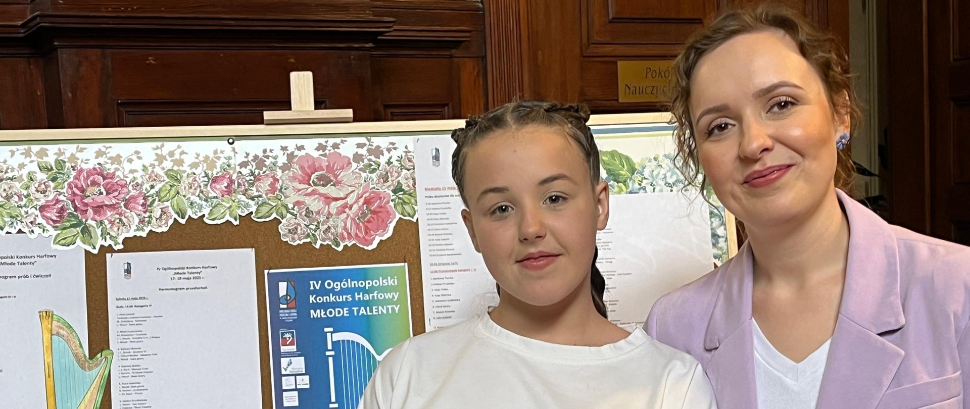 Uczennica i nauczycielka na tle tablicy z informacjami konkursowymi.