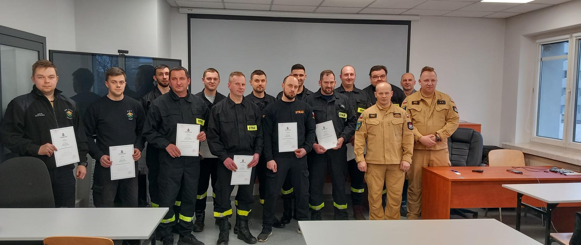 Strażacy jednostek ochotniczych straży pożarnych oraz dwóch strażaków Państwowej Straży Pożarnej stoją do wspólnego zdjęcia po wręczeniu zaświadczeń o zdanym egzaminie naczelników OSP. Strażacy OSP w umundurowaniu koszarowym a strażacy PSP w umundurowaniu służbowym. Zdjęcie na sali wykładowej Ośrodka Szkolenia KW PSP w Opolu.