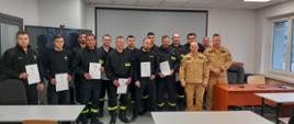 Strażacy jednostek ochotniczych straży pożarnych oraz dwóch strażaków Państwowej Straży Pożarnej stoją do wspólnego zdjęcia po wręczeniu zaświadczeń o zdanym egzaminie naczelników OSP. Strażacy OSP w umundurowaniu koszarowym a strażacy PSP w umundurowaniu służbowym. Zdjęcie na sali wykładowej Ośrodka Szkolenia KW PSP w Opolu.