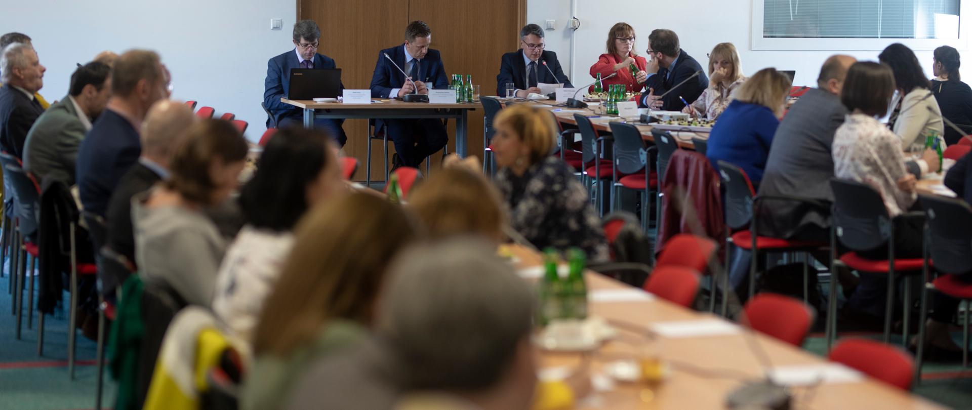 W sali przy długich stołach, na których stoją mikrofony, siedzą uczestnicy posiedzenia.