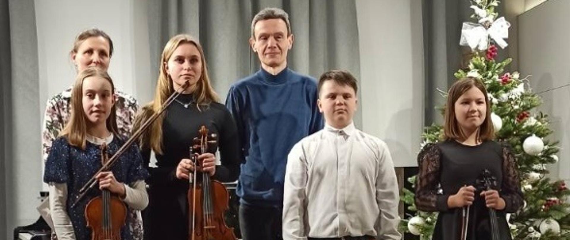 Zdjęcie przedstawia uczniów ze skrzypcami i nauczycieli i stojących na scenie w jednym rzędzie. W tle widać częściowo zasłonięte kotary w szarym kolorze. Z prawej strony za uczniami stoi ubrana choinka.