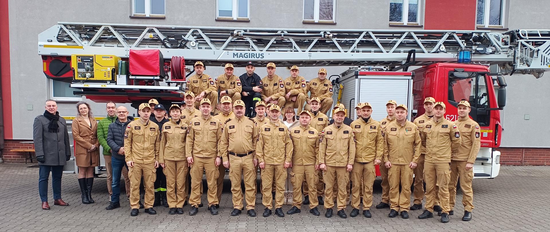 Na zdjęciu widać dużą grupę strażaków pozujących do pamiątkowej fotografii przed nowoczesnym wozem strażackim marki Magirus. Większość osób ubrana jest w piaskowe mundury służbowe z charakterystycznymi czapkami z daszkiem, natomiast kilka osób wyróżnia się ciemnymi, czarnymi strojami oraz ubiorem cywilnym. Zespół stoi na wybrukowanym placu przed budynkiem jednostki, a niektórzy strażacy zajęli miejsca wyżej, siedząc na elementach konstrukcyjnych drabiny mechanicznej.