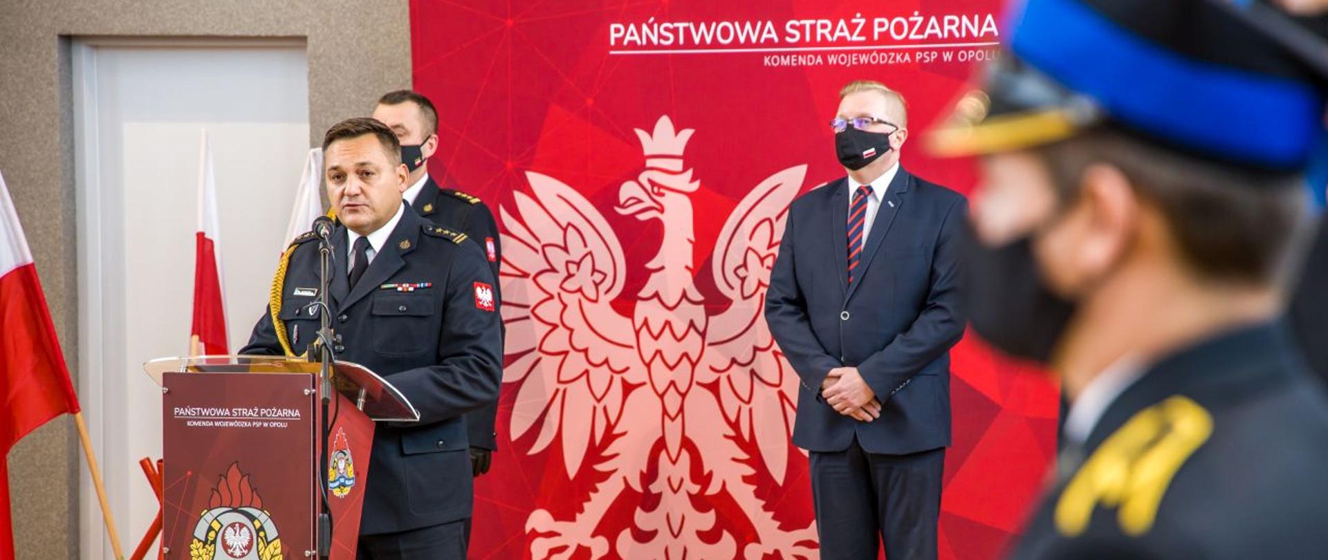 Opolski Komendant Wojewódzki PSP w Opolu st. bryg. Krzysztof Kędryk przemawia przy mównicy podczas uroczystej zbiórki w sali gimnastycznej KW PSP w Opolu. W drugim rzędzie stoi Wicewojewoda Opolski oraz Zastępca Opolskiego Komendanta Wojewódzkiego PSP w Opolu.