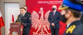 Opolski Komendant Wojewódzki PSP w Opolu st. bryg. Krzysztof Kędryk przemawia przy mównicy podczas uroczystej zbiórki w sali gimnastycznej KW PSP w Opolu. W drugim rzędzie stoi Wicewojewoda Opolski oraz Zastępca Opolskiego Komendanta Wojewódzkiego PSP w Opolu.