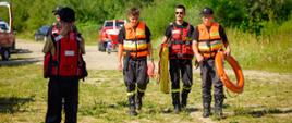 Ujęcie od przodu przedstawiające trzech strażaków idących trawiastą ścieżką po piaszczystym terenie. Wszyscy mają na sobie pomarańczowe kamizelki asekuracyjne. Strażak po prawej niesie pomarańczowe koło ratunkowe, a pozostali sprzęt ratowniczy w rękach. W tle widoczne są krzewy i samochód terenowy.