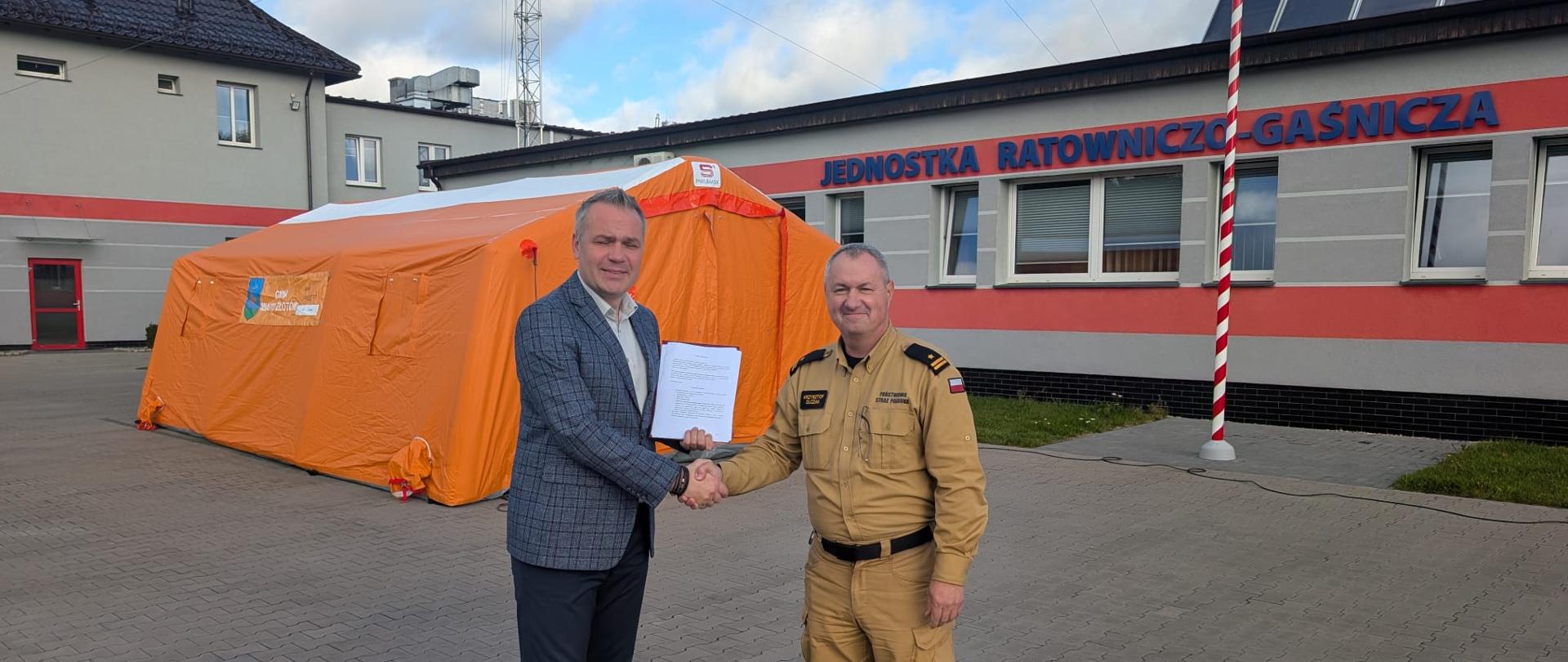 Na placu przed budynkiem jednostki ratowniczo-gaśniczej dwóch mężczyzn uściskiem dłoni symbolizuje przekazanie dokumentu. Jeden z nich ma na sobie marynarkę i trzyma kartkę, drugi ubrany jest w mundur strażacki. W tle znajduje się budynek jednostki z napisem „JEDNOSTKA RATOWNICZO-GAŚNICZA”, maszt oraz duży pomarańczowy namiot ratowniczy. Nad budynkiem widoczne są panele fotowoltaiczne, a w tle częściowo zachmurzone niebo.