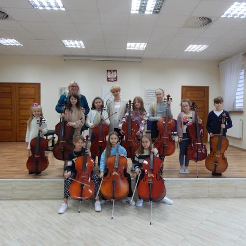 Fotografia przedstawia uczniów klasy wiolonczeli stojących z wiolonczelami oraz Panią Annę Skibińską i Urszulę Właźniak, które stoją razem z nimi. W tle brązowe drzwi, na górze wisi Godło Polski, w prawym rogu widać okna i białe firanki. 