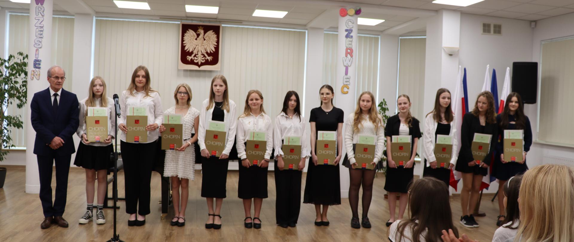 Zakończenie roku szkolnego. Dyrektor oraz uczniowie szkoły muzycznej stoją w sali. Wszyscy pozują do zdjęcia. Dzieci ubrane są odświętnie w białe koszule oraz czarne spodnie lub spódnice. W rękach trzymają dyplomy i książki. Mężczyzna ubrany jest w granatowy garnitur.