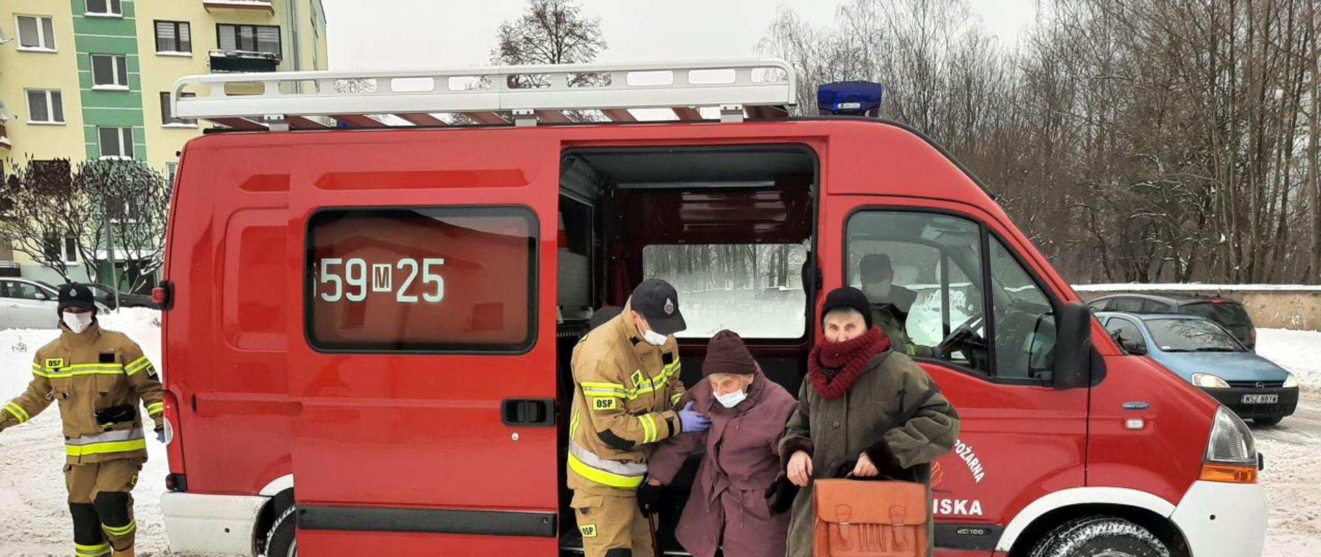 Fotografia przedstawiająca strażaków z OSP Chlewiska pomagających wysiąść starszej kobiecie z samochodu strażackiego bus celem dowozu do SPZOZ Chlewiska na szczepienie przeciw COVID-19 w ramach Narodowego Programu Szczepień.