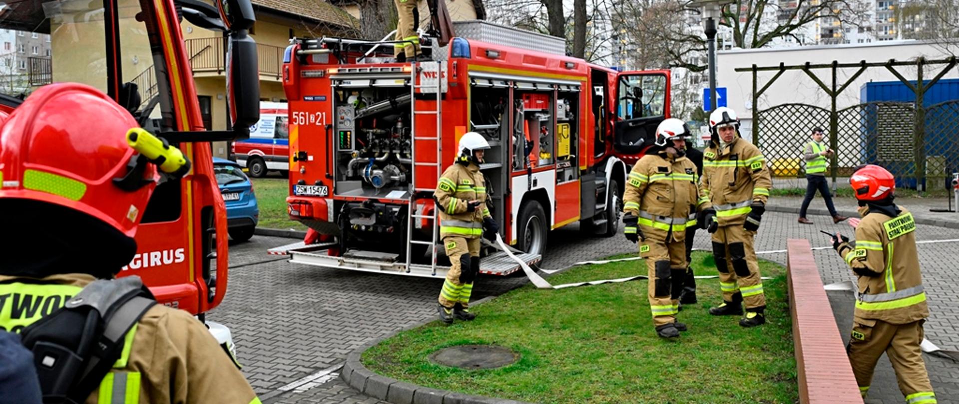 Zdjęcie przedstawia trzech strażaków. Strażacy w czerwonych hełmach, piaskowych oraz ciemnych mundurach. Strażacy wyciągają sprzęt ratowniczy z pojazdu pożarniczego. W tle samochód pożarniczy koloru czerwonego.