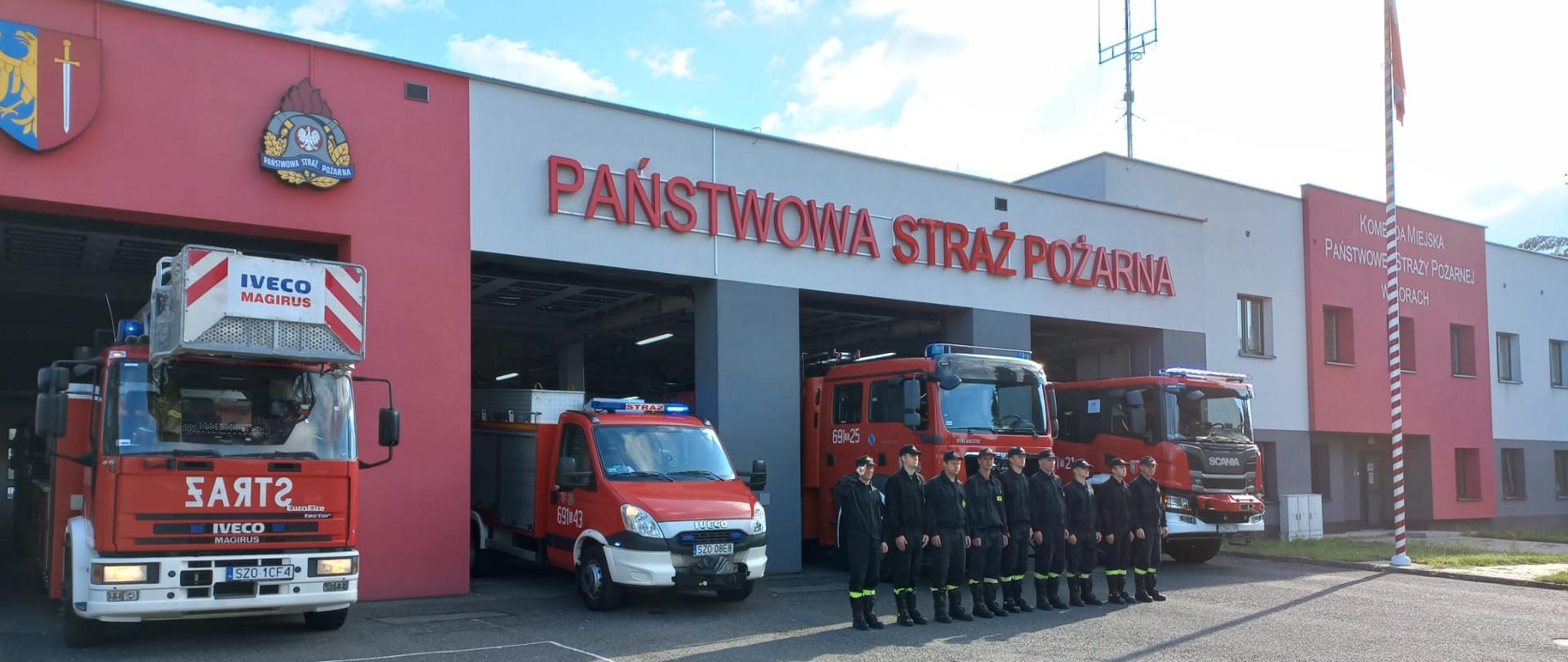 Strażacy stoją w dwuszeregu na tle samochodów pożarniczych i budynku Komendy. Pierwszy z nich salutuje.