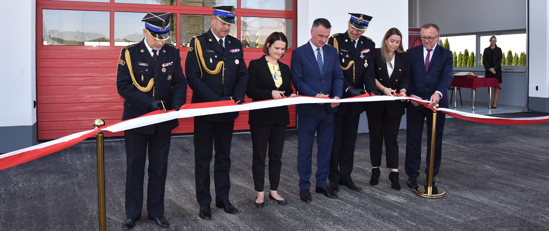 Na zdjęciu widzimy moment przecięcia wstęgi przez zaproszonych gości, a także kierownictwo komendy powiatowej i wojewódzkiej. Za przecinającymi wstęgę osobami znajduje się rozbudowana część obiektu strażnicy.