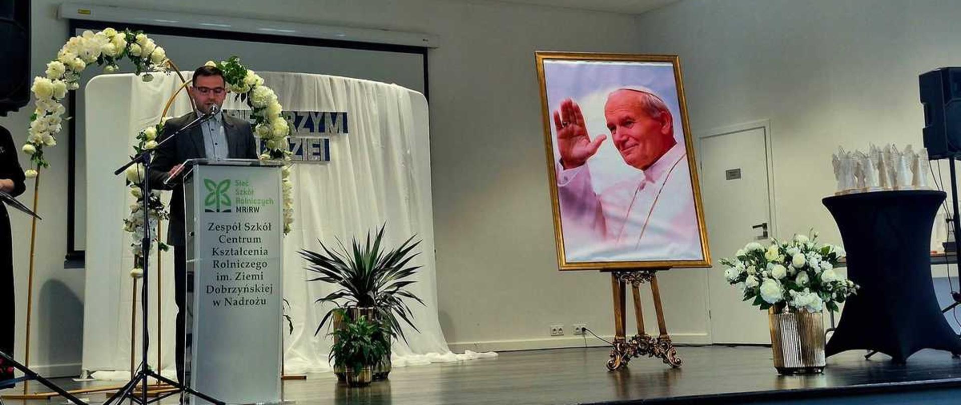 To zdjęcie przedstawia uroczystość odbywającą się w placówce edukacyjnej należącej do sieci Zespołów Szkół Centrum Kształcenia Rolniczego (nadzorowanych przez Ministerstwo Rolnictwa i Rozwoju Wsi).
Na mównicy widoczne jest logo oraz nazwa Zespołu Szkół Centrum Kształcenia Rolniczego.
Wydarzenie odbywa się w sali udekorowanej kwiatami.
Obok mównicy znajduje się portret przedstawiający Jana Pawła II.
Mężczyzna stojący za mównicą wygłasza przemówienie w obecności zebranych.
