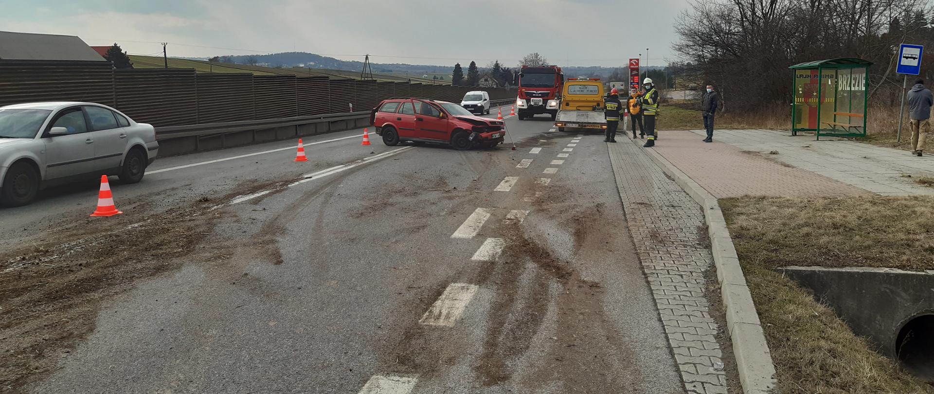 Na zdjęciu widoczny samochód osobowy na środku jezdni. W oddali widoczny samochód pożarniczy i dwóch strażaków. Po prawej stronie przystanek autobusowy. Jezdnia zabrudzona ziemią i sorbentem do usuwanie plam substancji oleistych.