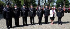 Zdjęcie zrobione na zewnątrz podczas uroczystości. W szeregu stoją strażacy ochotnicy, wojewoda podkarpacki i oficer PSP. W tle stoją ustawieni pozostali uczestnicy uroczystości.