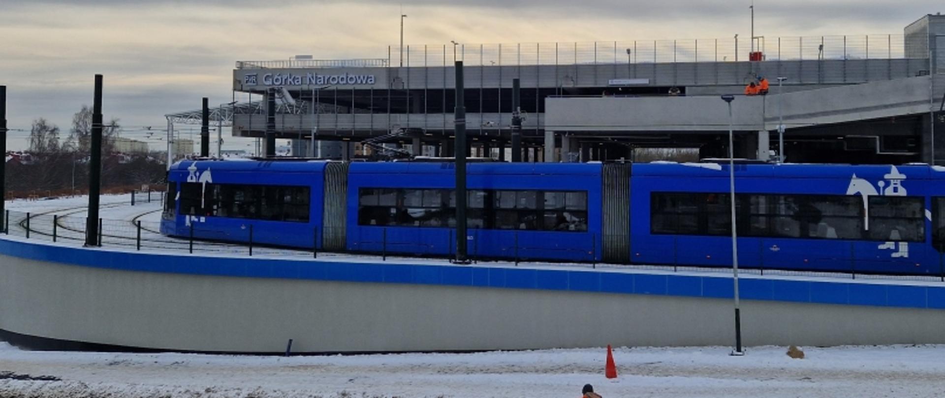 Niebieski tramwaj na nowo otwartym węźle przesiadkowym Górka Narodowa w Krakowie. Widoczne podwyższone torowisko, budynek stacji z napisem „Górka Narodowa” oraz śnieg na ziemi.