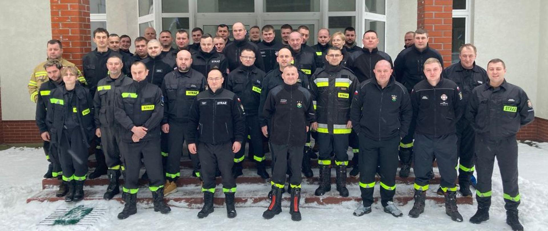 Zdjęcie wykonane przed budynkiem ośrodka szkolenia. Na pierwszym planie stoją uczestnicy kursu. Strażacy w czarnych ubraniach koszarowych.