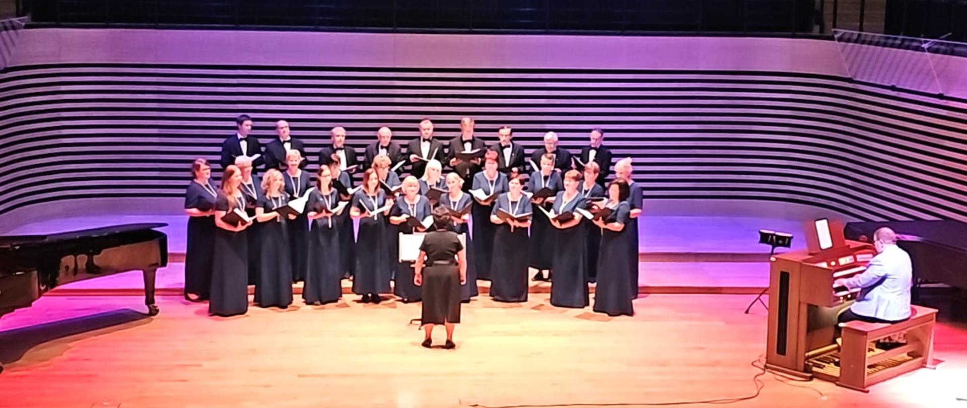 19 kobiet stoi w dwóch rzędach, za nimi na podwyższeniu w rzędzie stoi 9 mężczyzn, przed chórem tyłem do publiczności stoi kobieta, po prawej stronie na organach gra mężczyzna.