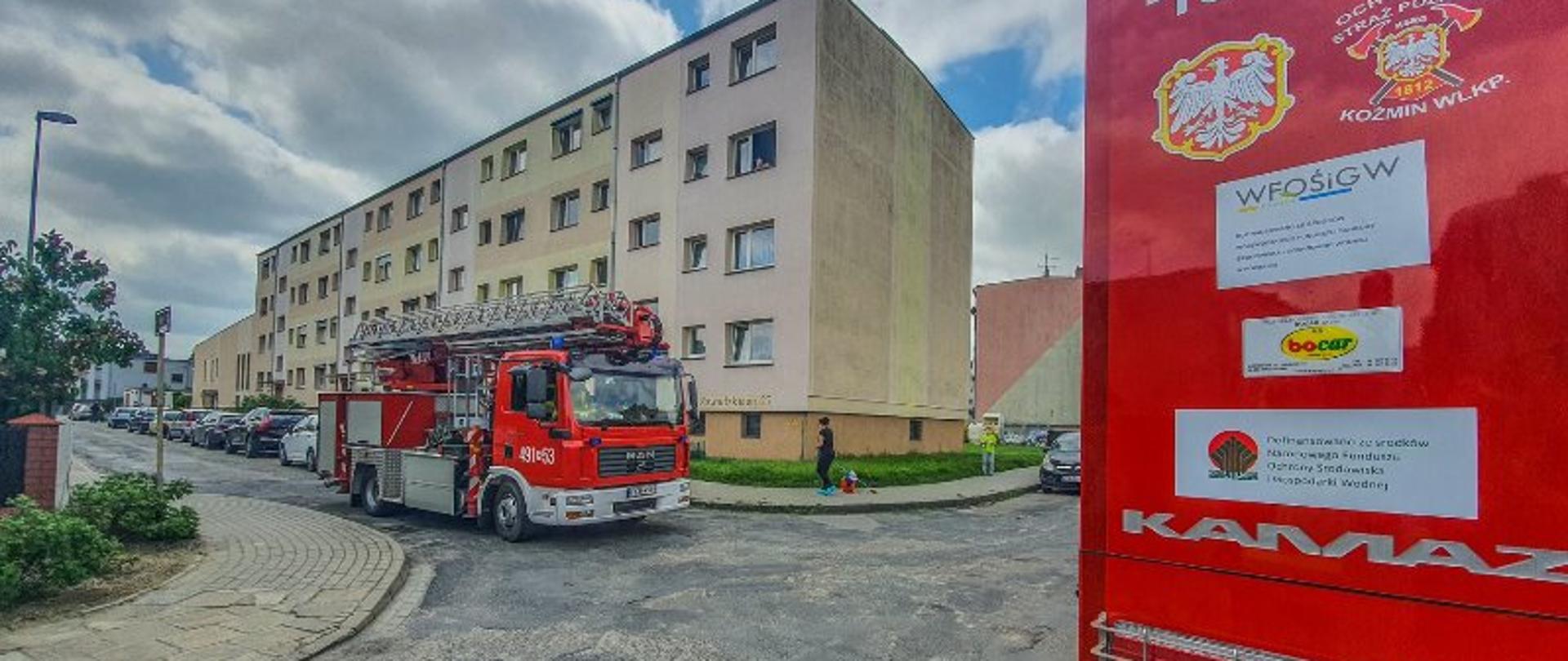 Przed budynkiem wielorodzinnym na drodze stoi samochód pożarniczy z Jednostki Ratowniczo - Gaśniczej. Przy budynku zaparkowane są samochody osobowe lokatorów. Ludzie poruszają się chodnikiem przy ulicy.