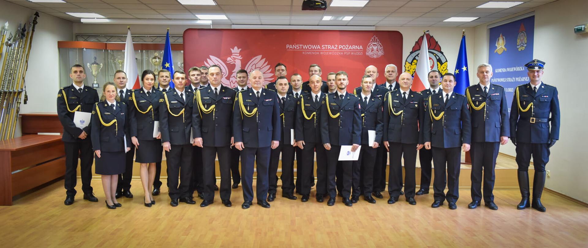 Duże grupowe zdjęcie oficerów Państwowej Straży Pożarnej (mężczyzn i kilku kobiet) w mundurach galowych, stojących razem po ceremonii. W tle duży transparent z logotypem PSP i napisem "PAŃSTWOWA STRAŻ POŻARNA KOMENDA WOJEWÓDZKA PSP W ŁODZI".