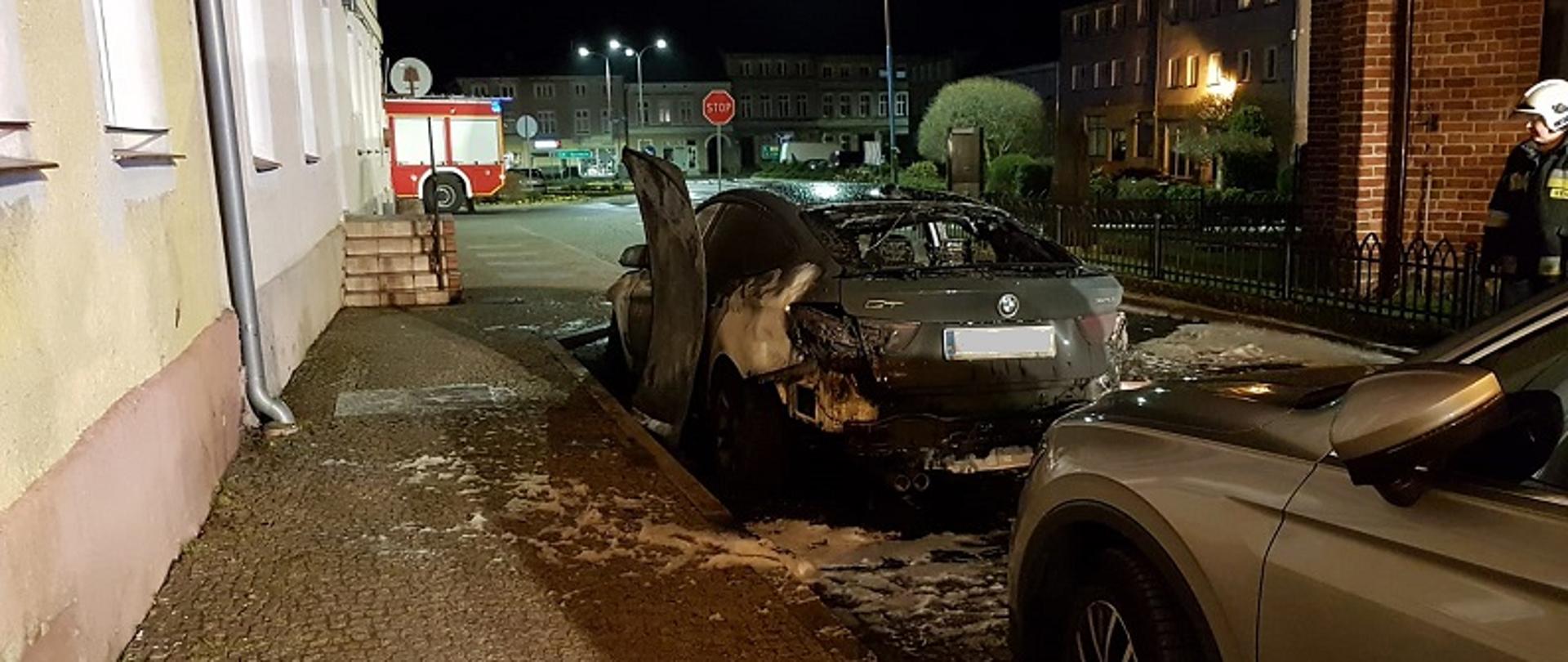 Zdjęcie przedstawia od widoczny od tyłu samochód osobowy BMW koloru szarego. Tylna szyba pojazdu jest wybita, a elementy nadwozia opalone. Auto stoi na drodze w perspektywie widoczne domy mieszkalne. Zdjęcie wykonane nocą