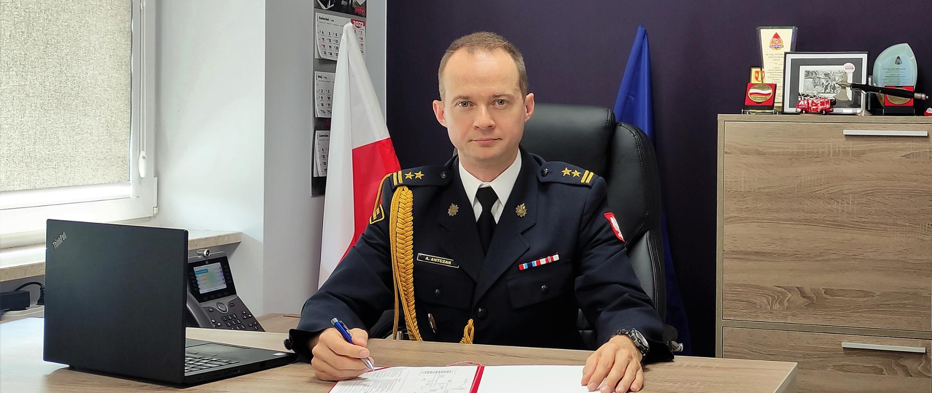 Zdjęcie przedstawia Komendanta Powiatowego PSP w Zgierzu, który przy swoim biurku podpisuję dokumenty. w tle widnieje komoda oraz fioletowa ściana z dwoma zdjęciami przedstawiającymi działania ratowniczo-gaśnicze.