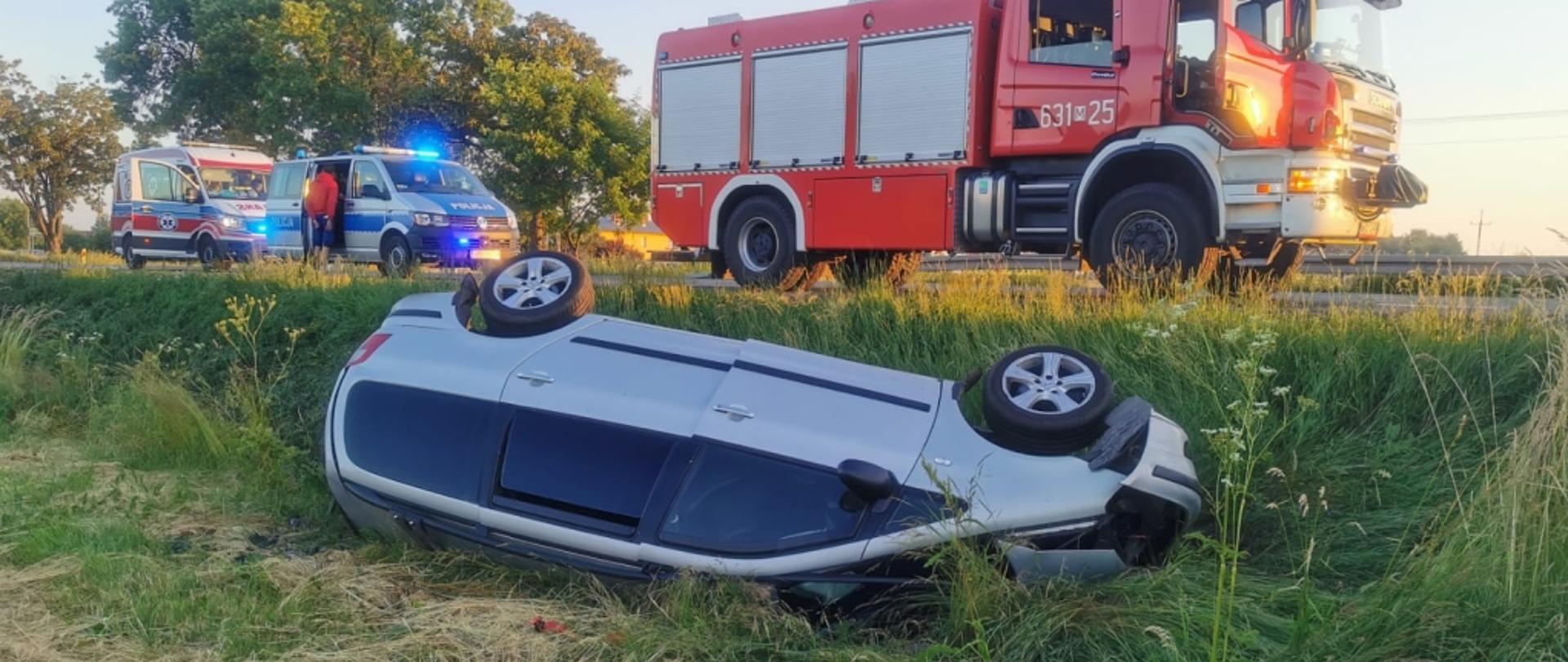 Zdjęcie przedstawia samochód osobowy leżący w przydrożnym rowie na dachu. Na drugim planie samochód straży pożarnej, radiowóz policji oraz ambulans.