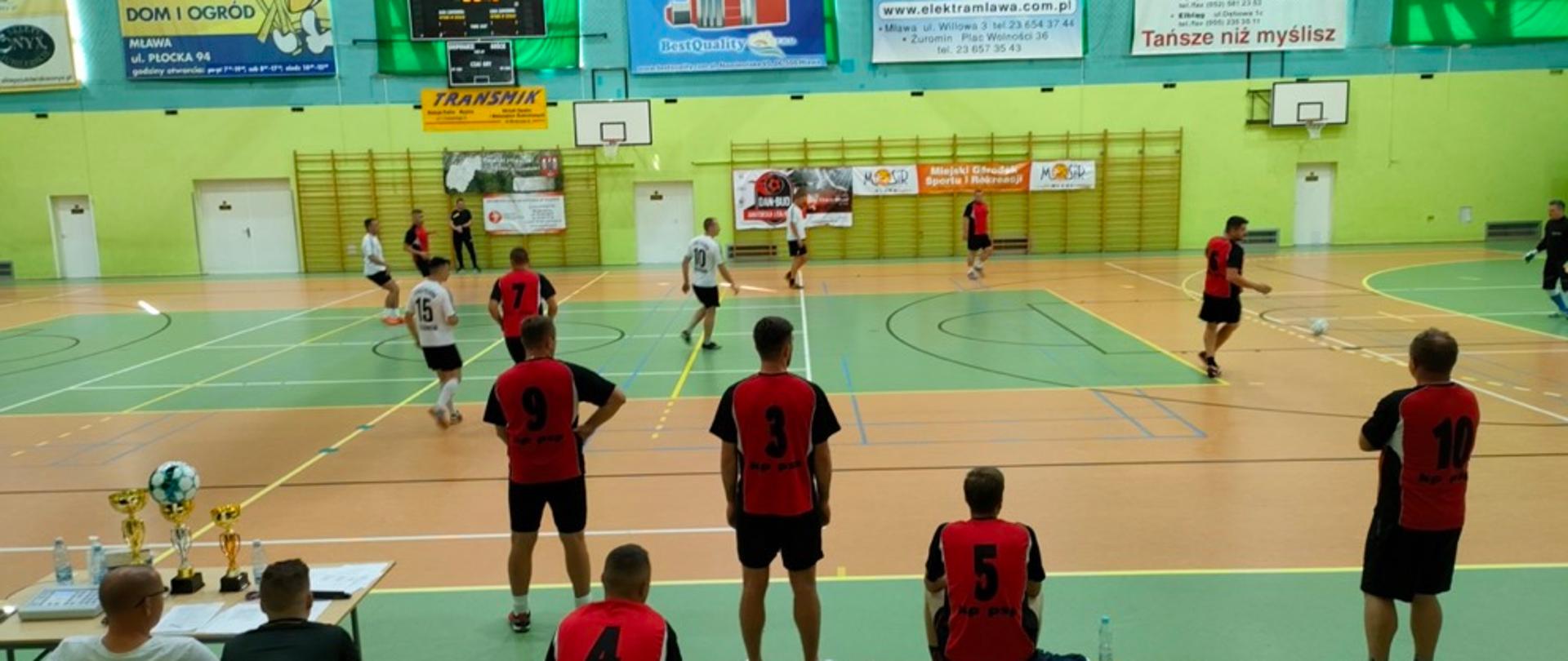 Sala sportowa, na której odbywa się mecz piłki nożnej. Na pierwszym planie siedzą kibice, w głębi jedna osoba kopie piłkę, a kilka osób asystuje. 